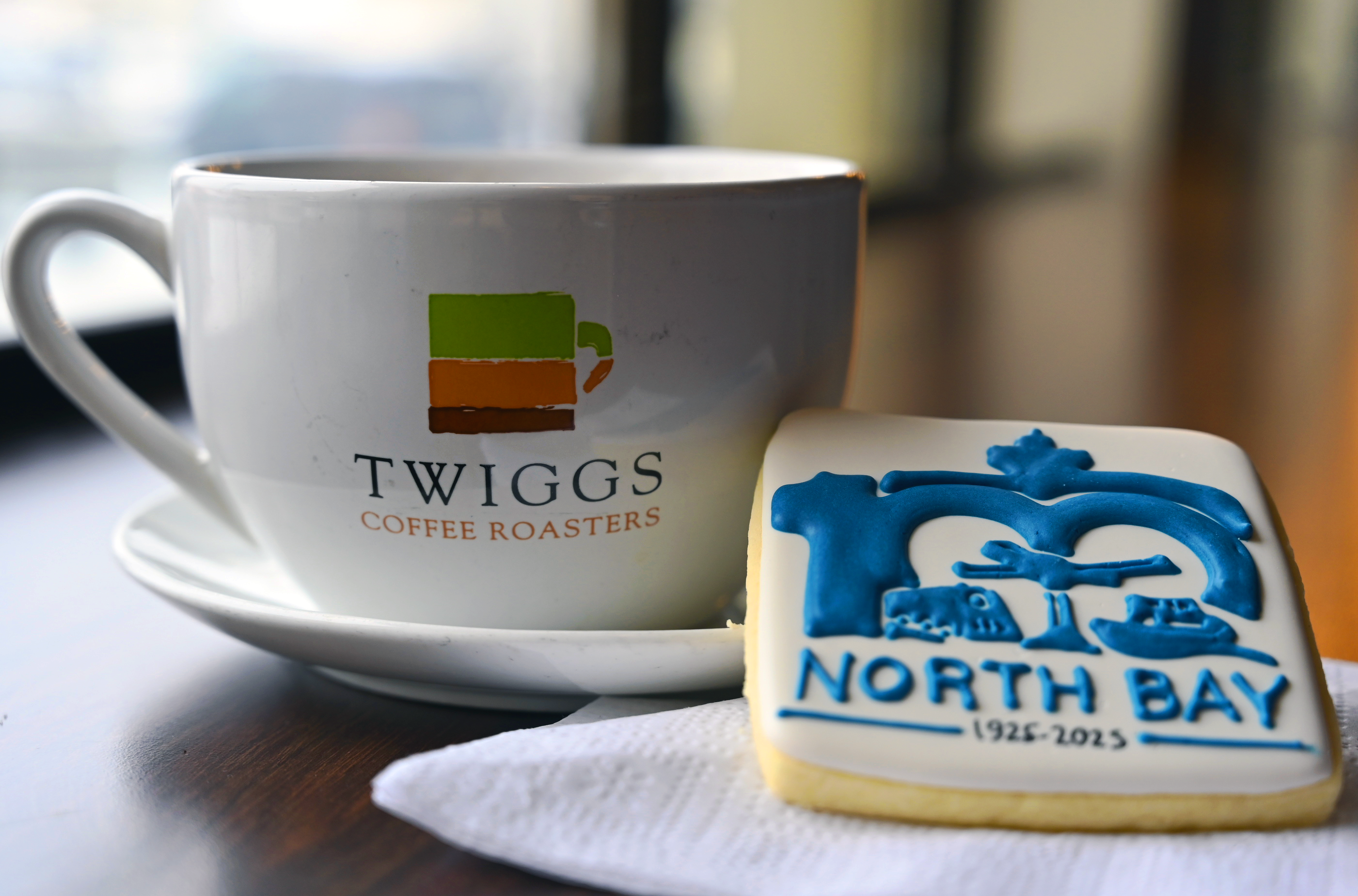 Twiggs branded white cup and saucer, next to it a square sugar cookie with white icing and North Bay Centennial logo in blue icing.